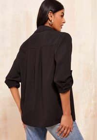 Black, lightweight blouse with rolled sleeves, back pleat detail, and a collar. Styled with blue jeans. Smooth texture, relaxed fit.