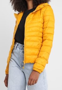 Żółta kurtka puchowa z kapturem, pikowana, z zamkiem błyskawicznym i elastycznymi mankietami, noszona na czarnej koszulce i jasnoniebieskich dżinsach.