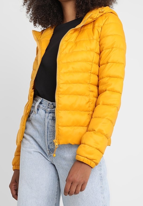 Żółta kurtka puchowa z kapturem, pikowana, z zamkiem błyskawicznym i elastycznymi mankietami, noszona na czarnej koszulce i jasnoniebieskich dżinsach.