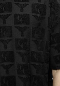 Μαύρο ύφασμα με ανάγλυφα υφασμένα γράμματα "Y" και "C" τοποθετημένα σε σκακιέρα σε ένα ρούχο με κοντά μανίκια.