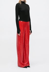 Personne portant un haut noir ajusté à manches longues et un pantalon rouge ample à sequins taille haute, debout sur un fond blanc uni.