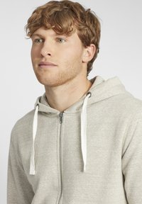 Lys grå zip-up hoodie lavet af blødt stof, med en justerbar hætte og hvide accenter. Glat tekstur med et afslappet design.