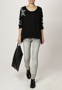 Un pull en tricot noir avec des accents d'étoiles argentées, porté avec un jean skinny gris clair et des bottines noires brillantes ; tenant un grand sac fourre-tout noir.