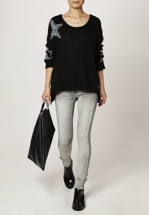 Un pull en tricot noir avec des accents d'étoiles argentées, porté avec un jean skinny gris clair et des bottines noires brillantes ; tenant un grand sac fourre-tout noir.