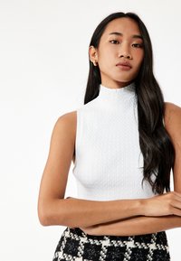 Vit, ärmlös polokrage topp med ribbad textur. Modellen har långt hår och bär en mönstrad svart-vit kjol.