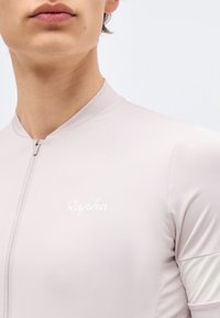 Κοντινό πλάνο ενός ατόμου που φοράει ανοιχτόχρωμη φανέλα ποδηλασίας Rapha με φερμουάρ μπροστά και αντανακλαστική λευκή ταινία στο μανίκι.