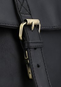 Primo piano di una borsa in pelle nera con una fibbia in ottone e hardware della tracolla, con cuciture visibili e una texture liscia.