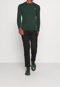 Homme portant un pull ras du cou vert foncé, un pantalon noir retroussé et des chaussures vertes à lacets, marchant devant un fond clair uni.