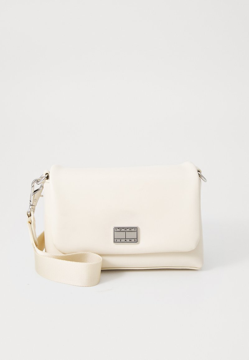 Bolso bandolera de imitación cuero color crema con una placa de logo en plata, correa ajustable, textura suave y diseño minimalista.