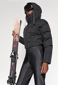 Must tänavapuhujaga must jakker, millel on kapuuts ja lukuga taskud, ning seljas on mustad suusipüksid. Isik hoiab käes graafilise disainiga suuski.