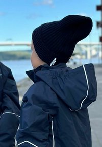 Svart ribbad stickad mössa med spetsig topp, bärs med en mörkblå vattenavvisande jacka med vita detaljer och huva.