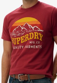 Rød bomulds t-shirt med grafik af bjerge og en solnedgang i orange og gul, med teksten "SUPERDRY MFG. CO. QUALITY GARMENTS" trykt med fed skrift.