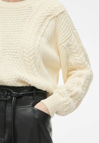 Pull en tricot crème avec motif en torsades sur les manches, associé à un pantalon en cuir noir avec une poche frontale et une ceinture.