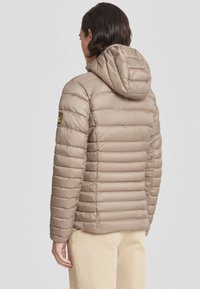 Persoon in een lichtbruine gewatteerde hoodie-jack, gezien van achteren, gecombineerd met beige broek tegen een effen achtergrond.