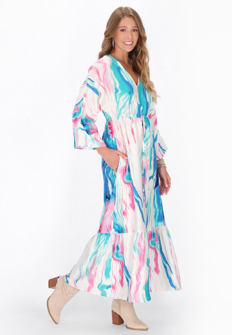 Mosolygó nő, hosszú, többszínű, forgó mintájú ruhát visel zsebekkel és bézs színű bokacsizmával, fehér háttér előtt pózol.