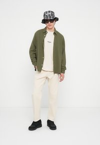 Groen overhemd over een beige T-shirt met het 'JJ Studios' logo, crèmekleurige broek, zwarte sneakers en een zwart-wit geruit bucket hat.