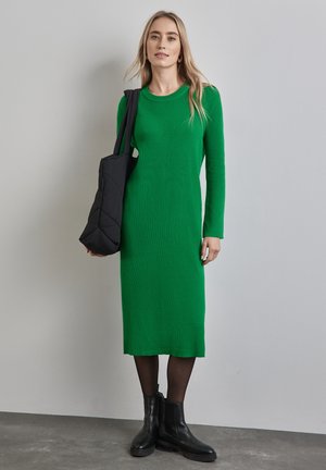 Robe longue verte à côtes avec des manches longues et un col rond. Associée à un sac fourre-tout matelassé noir et des bottines noires.