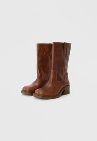 Bottines en cuir marron à bout rond, avec des languettes de tirage, des coutures discrètes et un petit talon carré. Texture lisse et design classique.