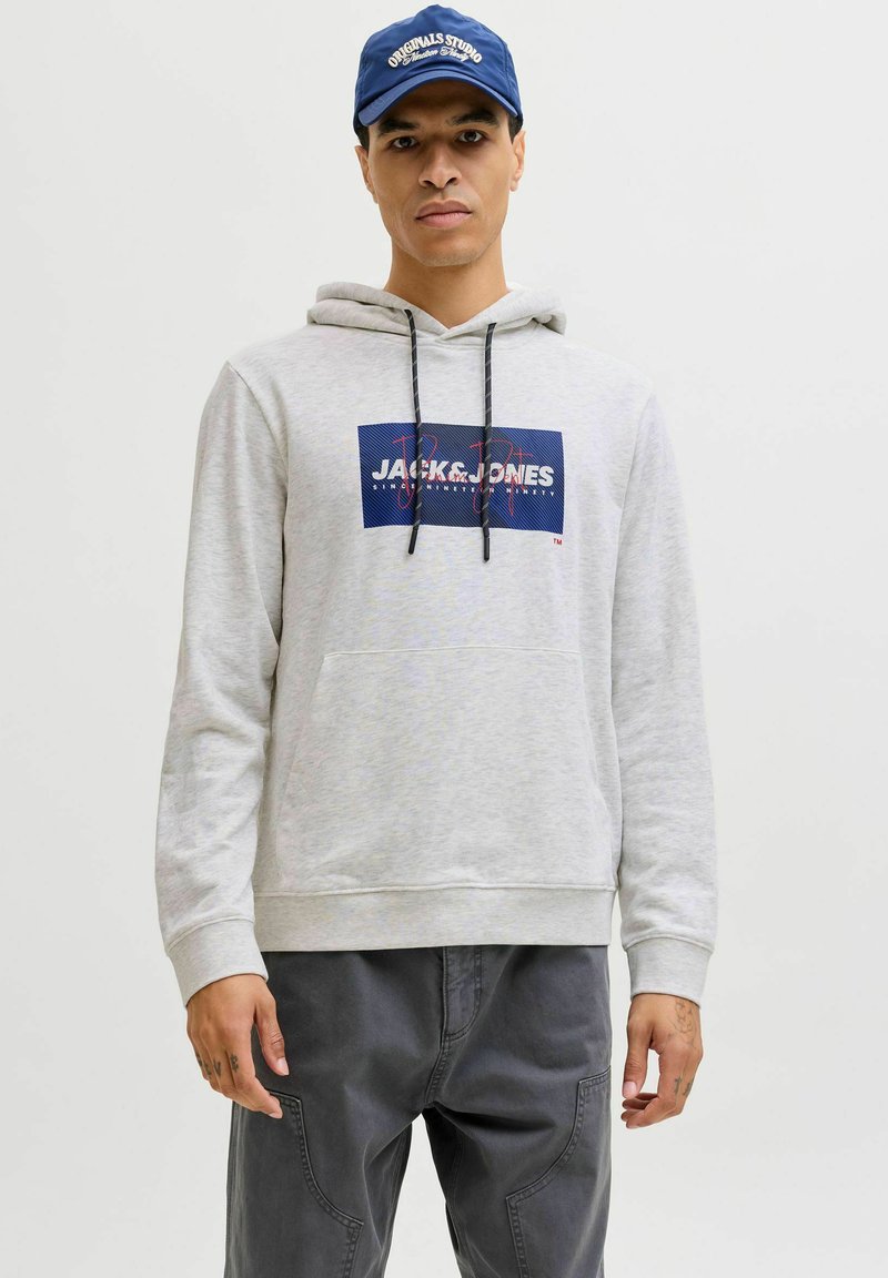 Homme portant un sweat à capuche gris clair Jack & Jones et une casquette bleue, debout devant un fond blanc uni, les mains détendues le long du corps.