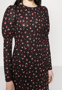 Kvinna iklädd en svart klänning med långa puffärmar och små blommönster i rött, vitt och rosa.