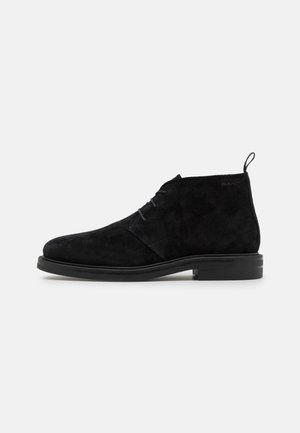 Schwarzer Wildleder-Stiefelette mit Schnürsenkeln, flachem Absatz und Zuglasche hinten, seitlich auf hellem einfarbigem Hintergrund dargestellt.