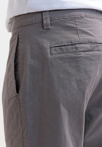 Close-up van een persoon die grijze broeken draagt met een zijzak en een achterzak met paspel, vergezeld van een wit shirt.