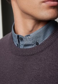 Pull en tricot violet foncé avec un col texturé bleu-gris arborant un motif géométrique et des boutons noirs. Gros plan sur la zone du cou.