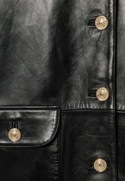 Giubbotto di pelle nera con bottoni dorati, texture liscia, con una tasca frontale e dettagli di cucitura visibili.