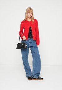 Red cardigan with gold buttons, black top, light blue wide-leg jeans, black handbag with curved handle, and black sneakers with brown soles.