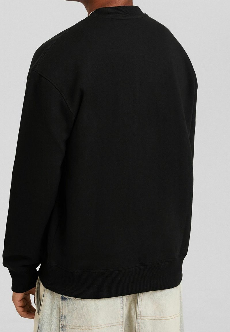 Homme portant un sweat-shirt noir uni à manches longues et un jean clair, vu de dos sur un fond uni.