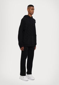 Felpa nera con motivo testurizzato, tasca a marsupio e cappuccio con coulisse, abbinata a pantaloni neri e sneakers bianche. Colore uniforme in tutta la componenente.