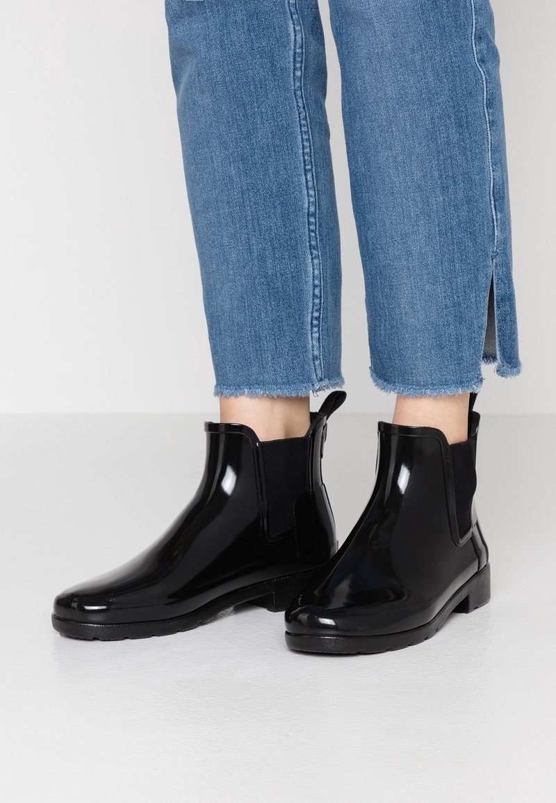 Bottes de pluie noires brillantes à la cheville portées avec un jean bleu raccourci aux ourlets effilochés et petites fentes latérales, debout sur un sol blanc.