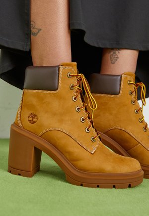 Tan suede ankle boots with brown collar, gold eyelets, and yellow laces. Chunky brown heel and logo detail on the side.