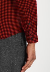 Camicia di flanella a quadri rossa e nera con polsini abbottonati, abbinata a pantaloni grigi con trama. La camicia ha un orlo curvo.