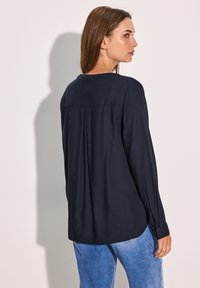 Blusa blu navy a maniche lunghe con una vestibilità comoda, caratterizzata da una piega sul retro, polsini con bottone e tessuto morbido e liscio. Abbinata a jeans azzurri chiari.