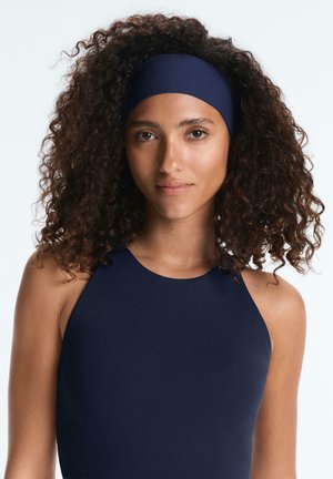 Donna con capelli ricci che indossa una canottiera sportiva blu navy e una fascia larga abbinata, rivolta in avanti con un'espressione neutra.