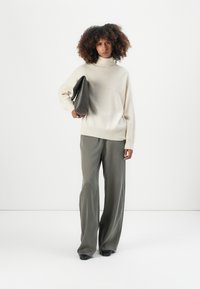 Beiger Rollkragenpullover und lockere olivgrüne Hose. Das Model hält eine dunkelgraue Clutch-Tasche, die eine glatte Textur und ein einfaches Design aufweist.