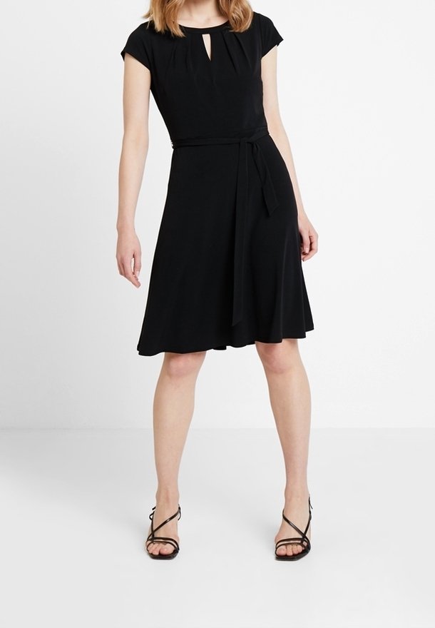 Femme portant une robe noire longueur genou avec une ceinture à la taille et un décolleté en forme de clé, associée à des sandales noires à talons hauts et brides.