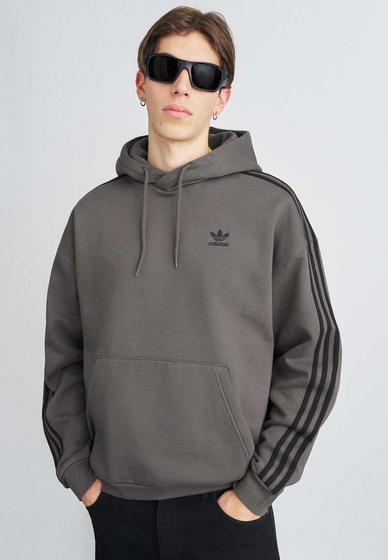 Pelēks kokvilnas kapuci ar priekšējo kabatu, melna Adidas logo un melni trīs svītru akcenti uz piedurknēm. Modeļa acīs ir lielas saulesbrilles.