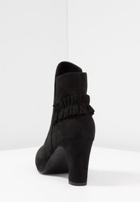 Černé semišové kotníkové boty s volánkovým akcentem na patě a zaoblenou špičkou. Patka má střední výšku a nabízí hladký, texturovaný povrch.