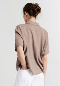 Camicia a maniche corte di colore marrone chiaro con un tessuto strutturato. Presenta un colletto e una vestibilità comoda, mostrata da dietro con una mano in tasca.