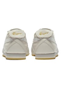 Bílé sportovní boty s gumovými podrážkami, koženými svršky a strukturovaným povrchem. Vyznačují se zlatým logem "Tiger" na patě a pruhovanými detaily.