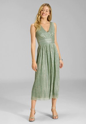 Młoda kobieta uśmiechająca się, ubrana w zieloną błyszczącą plisowaną sukienkę midi bez rękawów z dekoltem w serek i srebrne sandały na obcasie, stojąca na szarym tle.