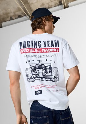 Mand iført hvid Red Bull Racing Team t-shirt med racerbils grafik og mørkeblå kasket, set bagfra mod ensfarvet baggrund.