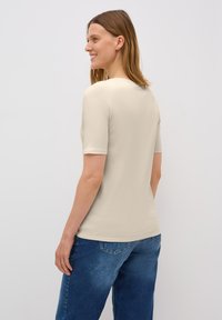 Světle béžové tričko s krátkým rukávem a kulatým výstřihem, vyrobené z měkkého materiálu. Kombinováno s volnými modrými džíny.