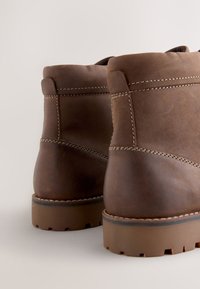 Hnedé kožené členkové čižmy s hrubými podrážkami, viditeľným prešívaním, zaoblenými hranami a hladkou textúrou, prezentujú jednoduchý dizajn.