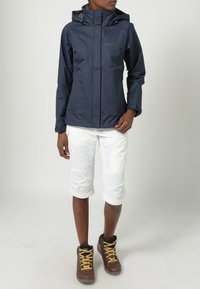Granatowa wodoodporna kurtka z przednim zamkiem, regulowanym kapturem i detalem z logo. W zestawie z białymi szortami do kolan i brązowymi butami trekkingowymi.
