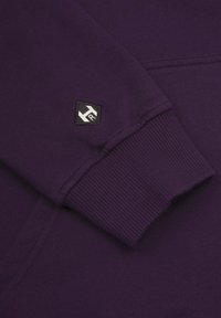 Lila sweatshirt med ribbad mudd och en liten svart logotagg med vita detaljer nära sömmen på ärmen. Slät textur. 