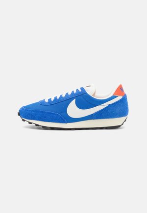 Niski, niebieski but sportowy Nike z białym logo Swoosh, białymi sznurowadłami, kremową podeszwą i pomarańczową metką na pięcie na białym tle.