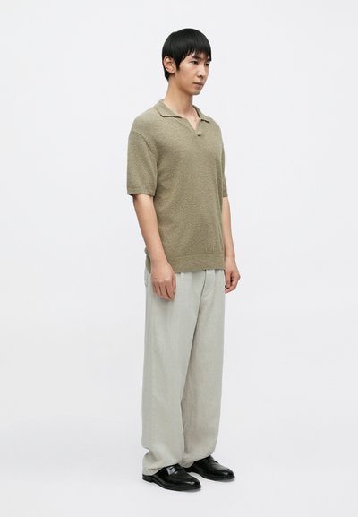 Πολό μπλούζα με κοντό μανίκι και υφή σε χακί χρώμα, συνδυασμένη με ανοιχτό γκρι παντελόνι φαρδιάς γραμμής και μαύρα παπούτσια, στέκεται μπροστά σε λευκό φόντο.